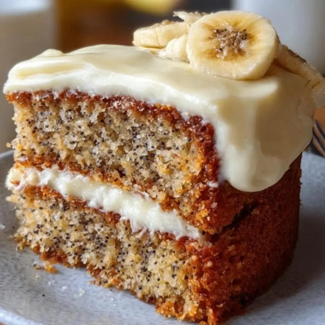 A close-up view of a slice of banana bread cake, showing the moist, dense crumb, thick layer of white cream cheese frosting, and a light dusting of cinnamon on top.