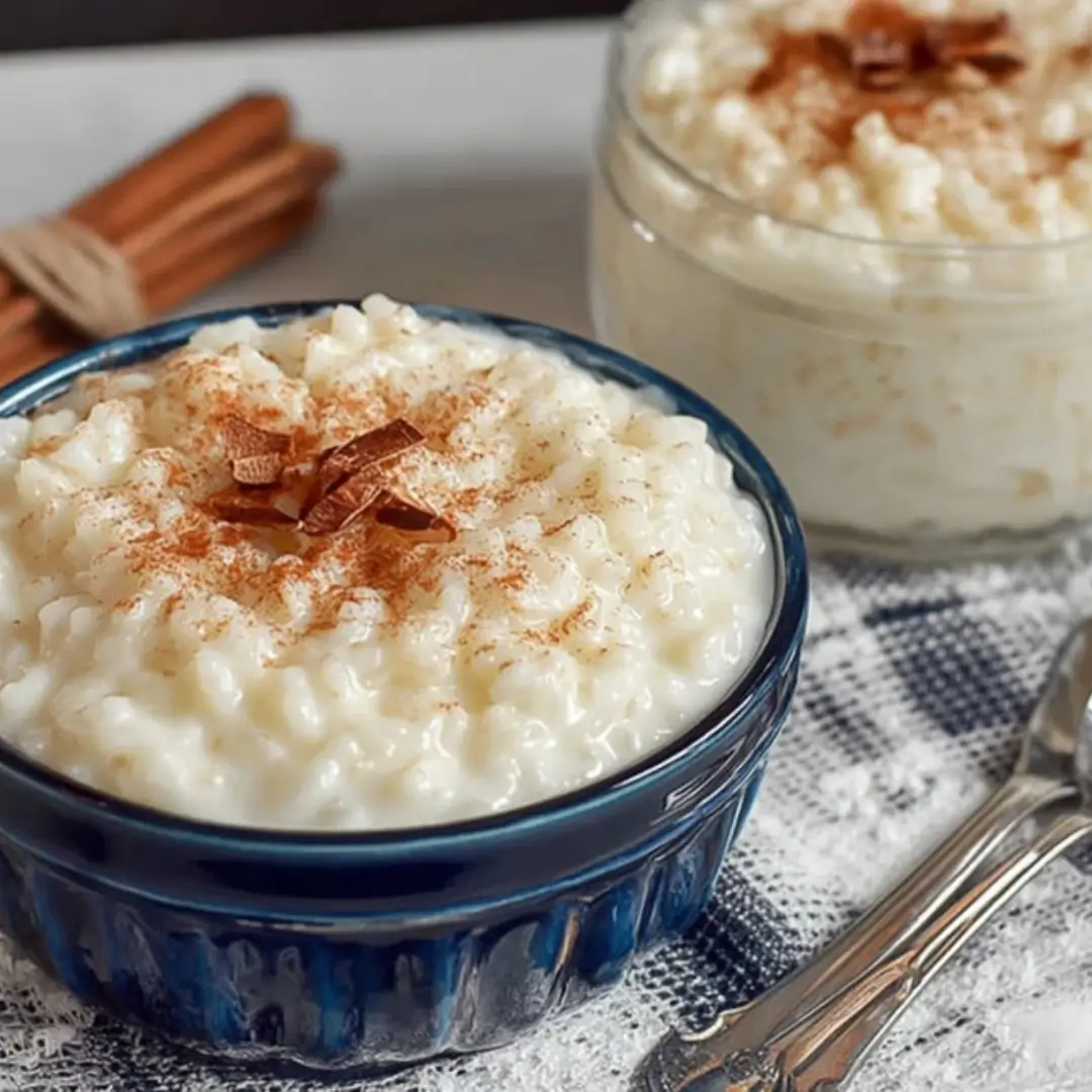 Creamy old-fashioned rice pudding dusted with cinnamon, served in a deep blue bowl with a jar of pudding and a spoon in the background.