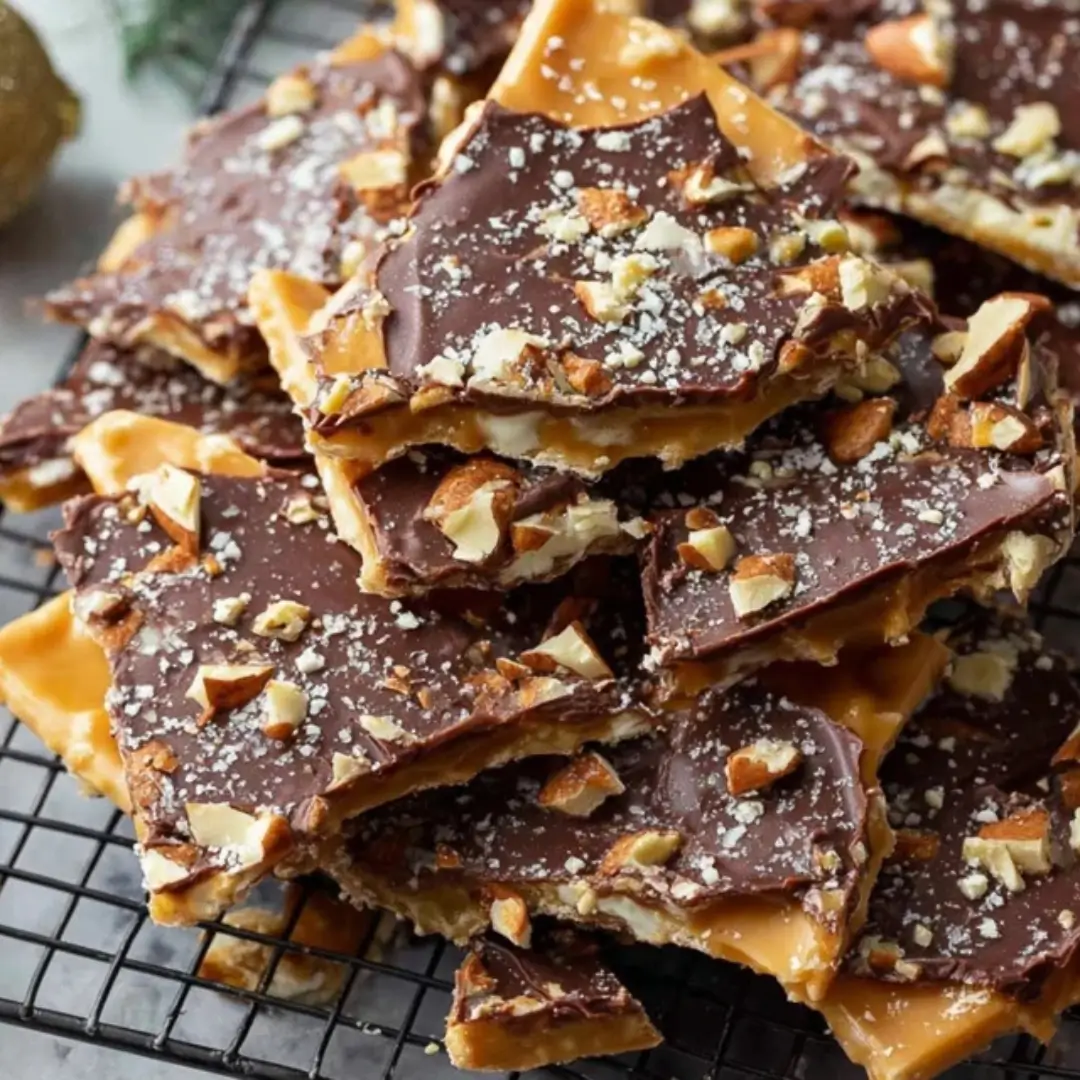 A stack of broken Christmas Crack toffee pieces on a black wire cooling rack. The toffee has a salty cracker base, a thick, golden caramel layer, and is topped with melted chocolate and chopped almonds or pecans, sprinkled with sea salt.
