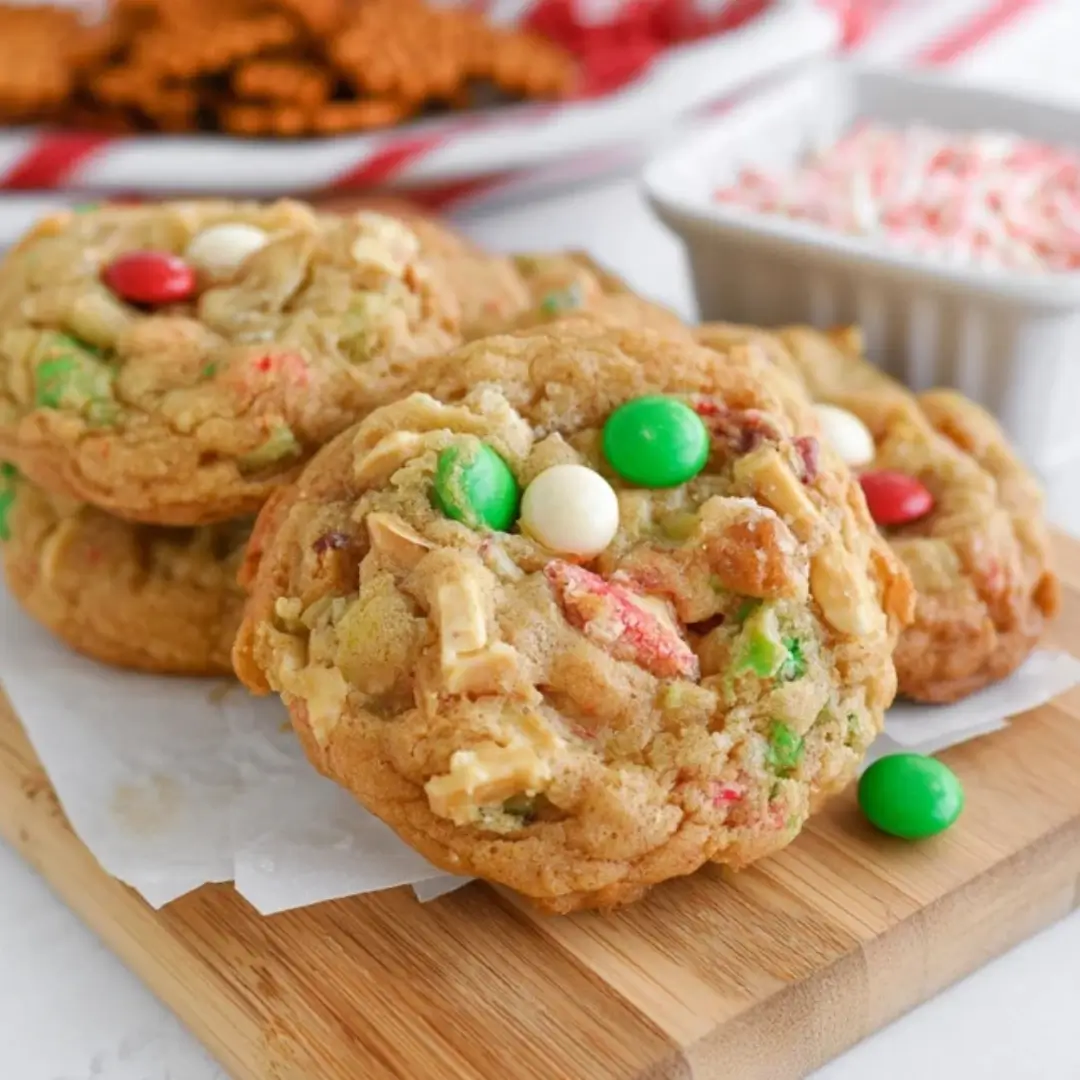 Stack of chewy Christmas kitchen sink cookies loaded with red & green M&M's, white chocolate chips, and crushed pretzels on a wooden board.