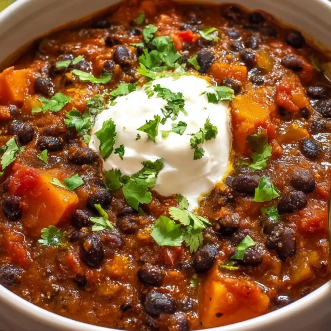 A close-up, top-down shot of a bowl filled with rich, dark Easy Black Bean Chili, featuring chunks of vegetables and black beans, topped with a dollop of white sour cream (or vegan cream) and garnished with fresh green cilantro.