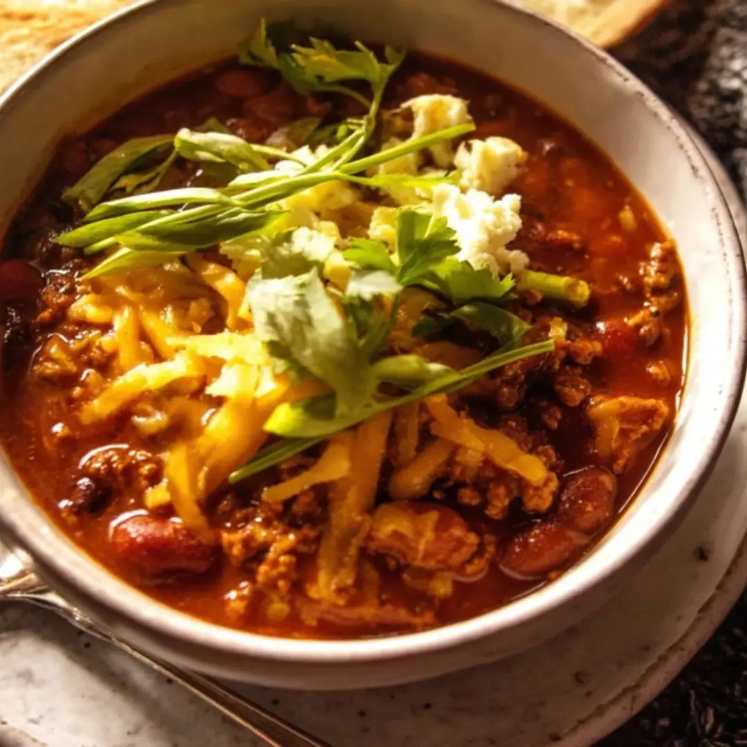 Bowl of rich, dark Easy Chipotle Cheddar Beef Chili topped with shredded cheddar cheese, sliced green onions, and fresh cilantro.