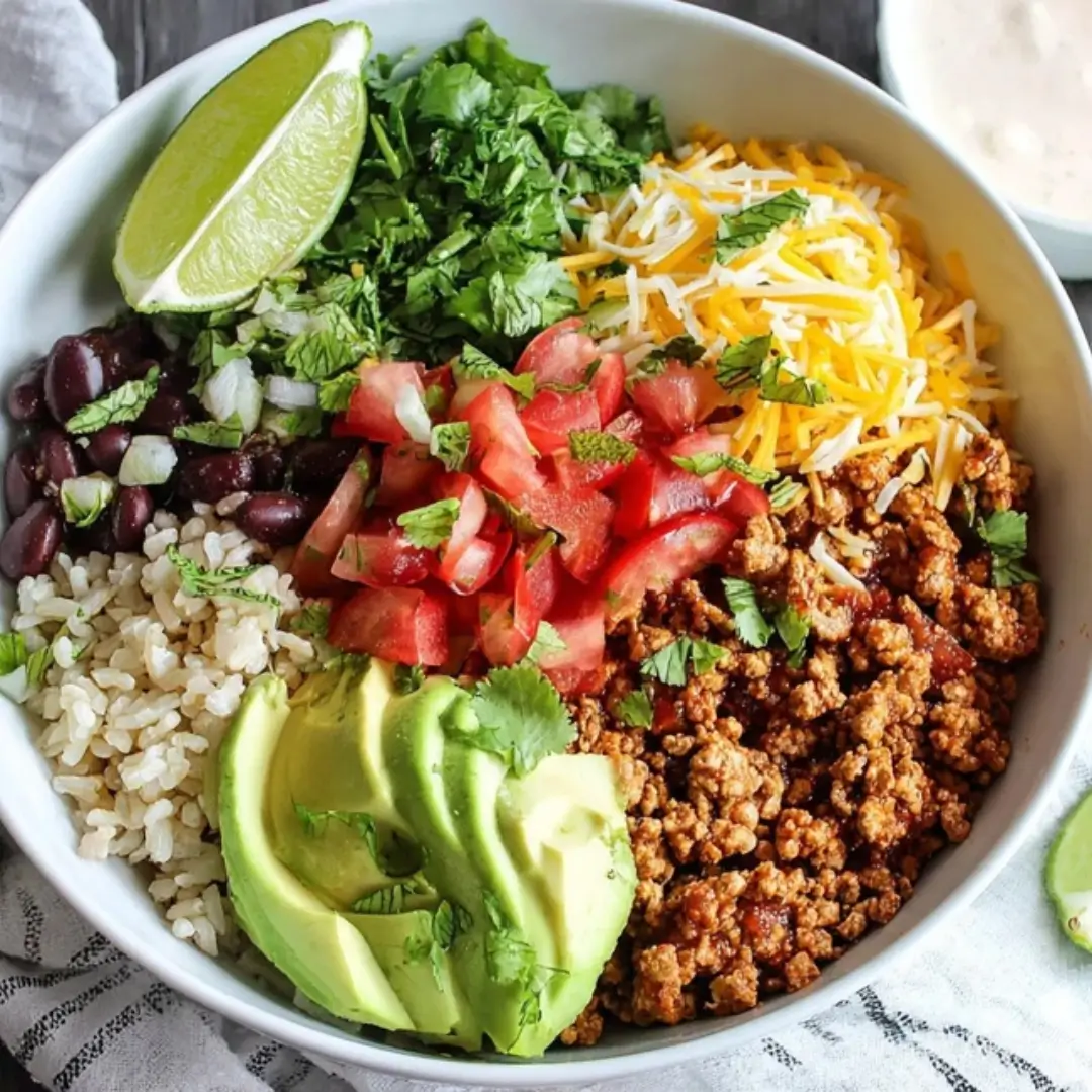 Close-up of a large white bowl filled with brown rice, seasoned ground taco meat (turkey or chicken), black beans, diced tomatoes, sliced avocado, shredded cheese, and fresh cilantro, garnished with a lime wedge.