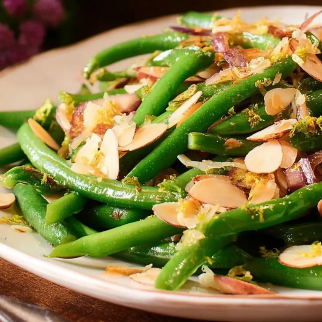A close-up of vibrant green beans almondine garnished with golden toasted sliced almonds, finely minced red onion (or shallots), and bright yellow lemon zest on a white plate.