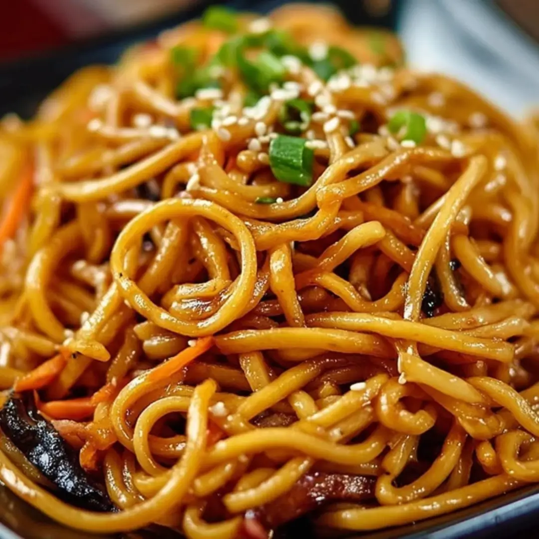 Close-up of glossy, savory Hibachi Noodles tossed with shredded carrots and green onions, garnished with sesame seeds and served in a black bowl.