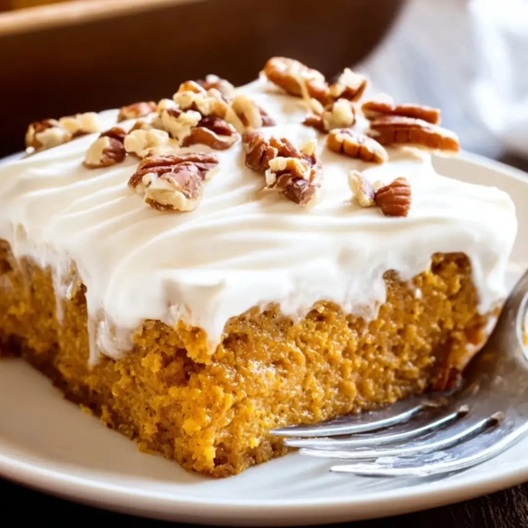 A slice of moist, spiced Pumpkin Poke Cake topped with a generous layer of white homemade whipped cream frosting and sprinkled with crushed pecans.