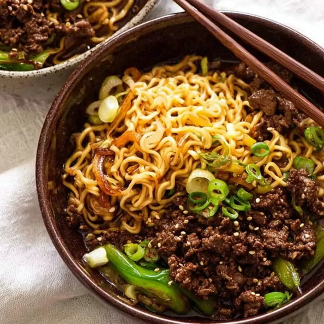 A close-up of a dark ceramic bowl filled with glossy ramen noodles and savory ground beef, garnished with sliced green onions and sesame seeds.