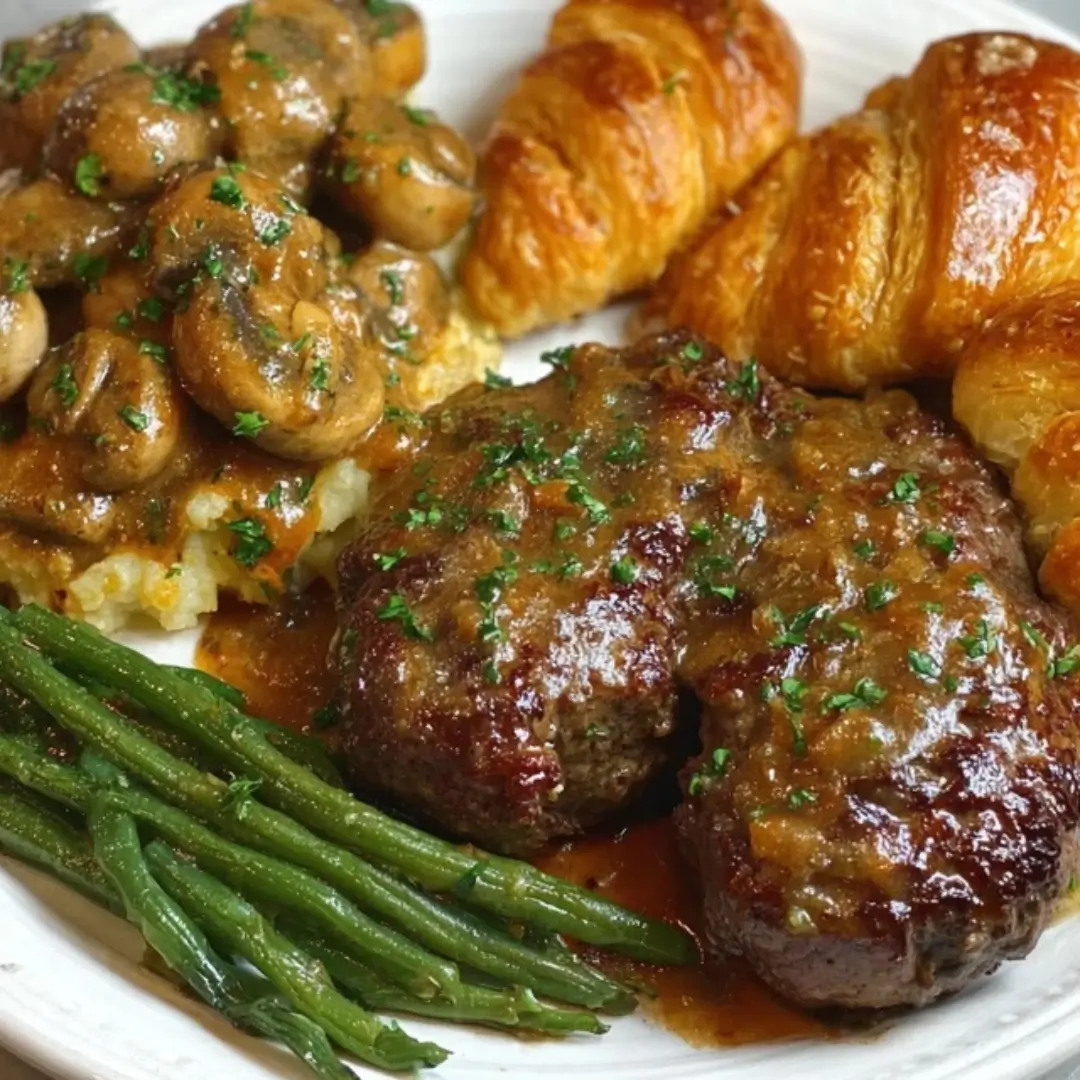 Two oval Salisbury steaks smothered in rich brown gravy, served alongside buttery mashed potatoes topped with sautéed mushrooms, a side of bright green beans, and golden honey butter croissants.