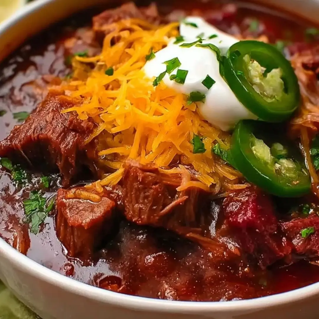 A close-up bowl of thick Beef Bacon Brisket Chili topped with shredded cheddar cheese, a dollop of sour cream, fresh jalapeño slices, and chopped cilantro.