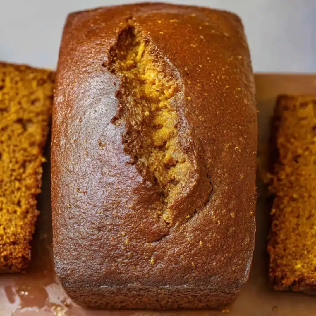 A top-down view of a golden brown, moist loaf of pumpkin bread with two thick slices cut on the sides, showing a soft, tender crumb.