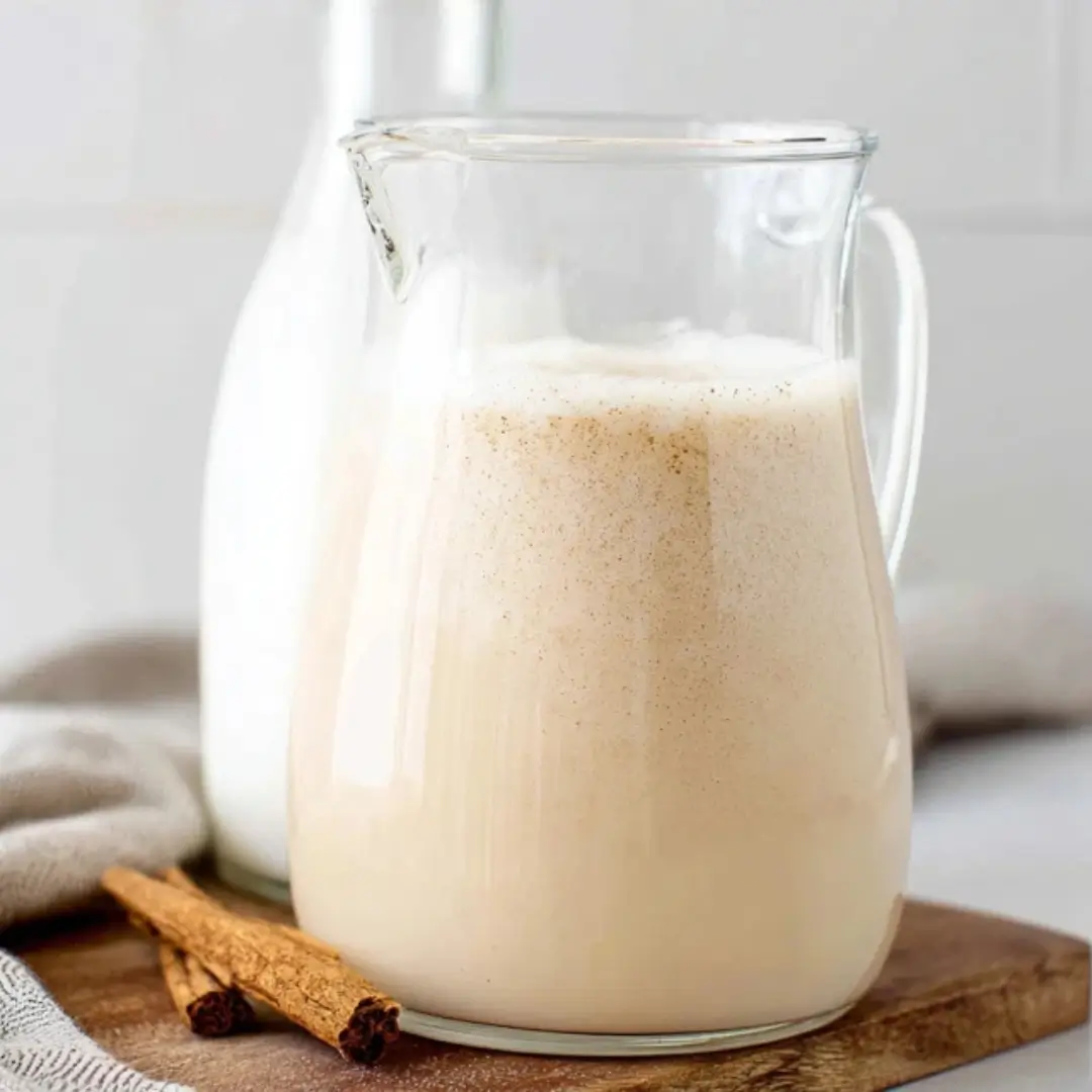 A glass pitcher filled with creamy, spiced non-alcoholic Rum Chata topped with ground cinnamon, sitting on a wooden board next to cinnamon sticks.