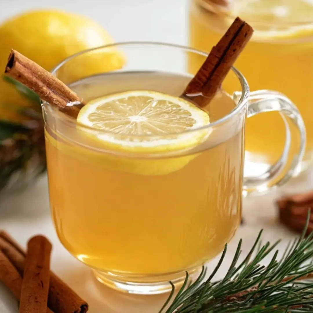 A glass mug of warm, golden non-alcoholic hot toddy garnished with a fresh lemon slice and a cinnamon stick, surrounded by festive pine needles and whole cinnamon.