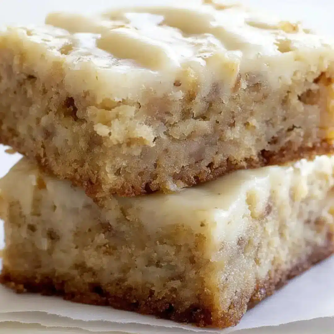 Two stacked thick-cut, moist banana bars with a creamy white frosting glaze, showing a dense and fudgy texture on parchment paper.