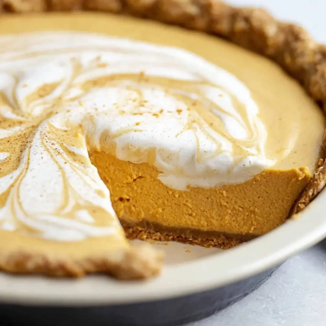 A close-up shot of a creamy pumpkin cream cheese pie in a flaky crust with a swirl of whipped cream topping and one slice missing to show the smooth texture.
