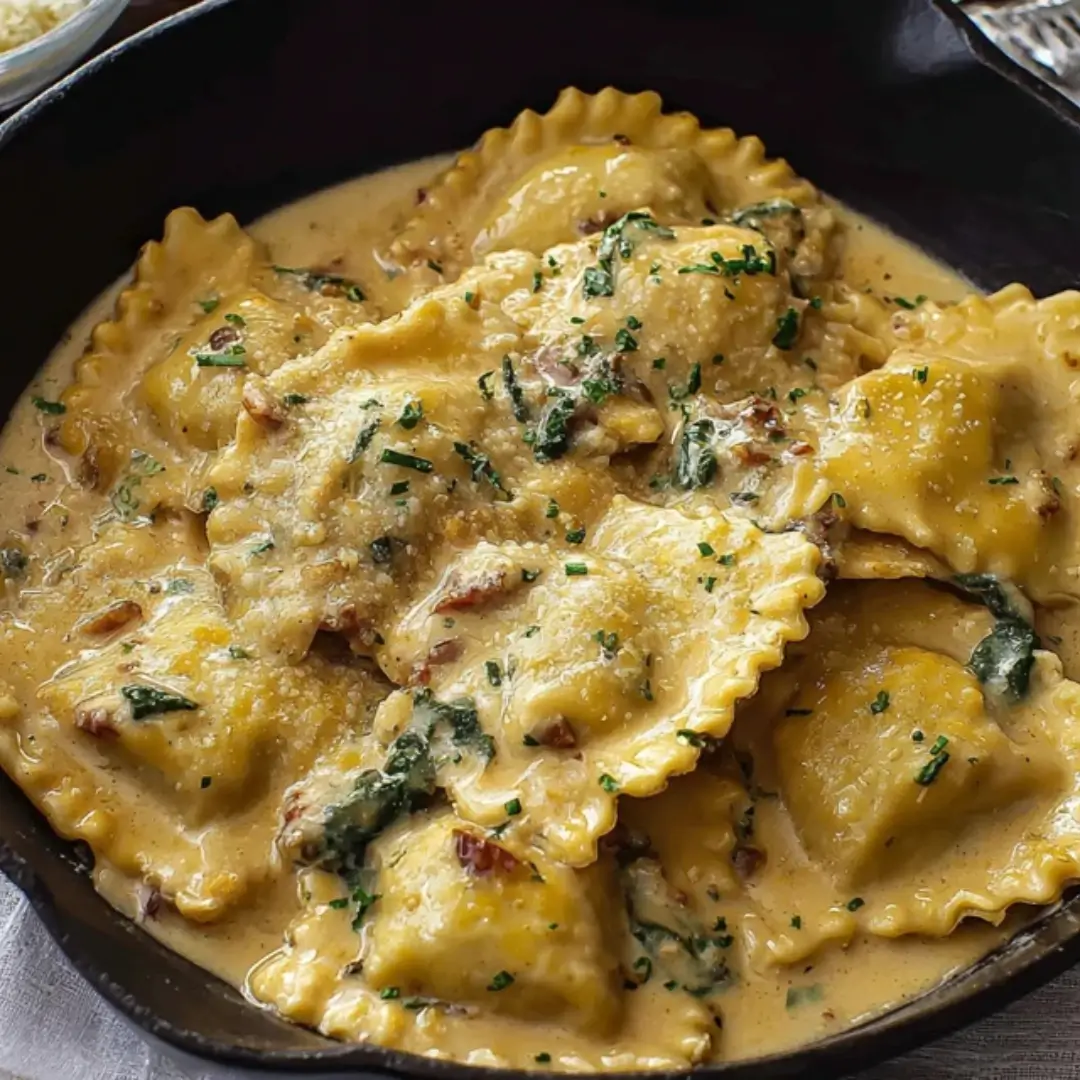 A close-up shot of golden cheese ravioli smothered in a creamy roasted garlic sauce with blistered cherry tomatoes and fresh herbs in a black cast-iron skillet.