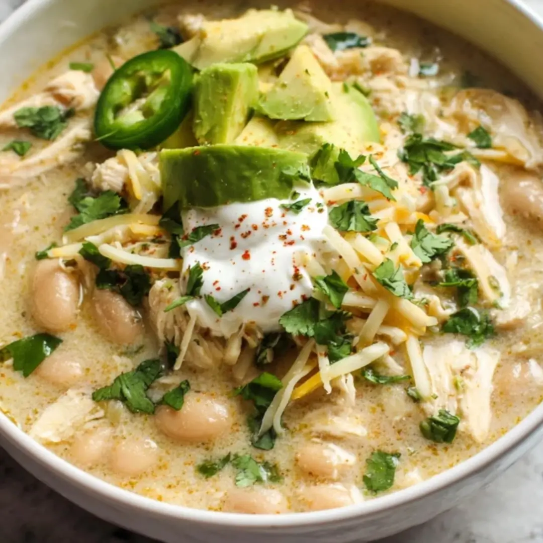 A close-up bowl of creamy white chicken chili topped with avocado slices, a dollop of sour cream, shredded cheese, fresh cilantro, and a slice of jalapeño.