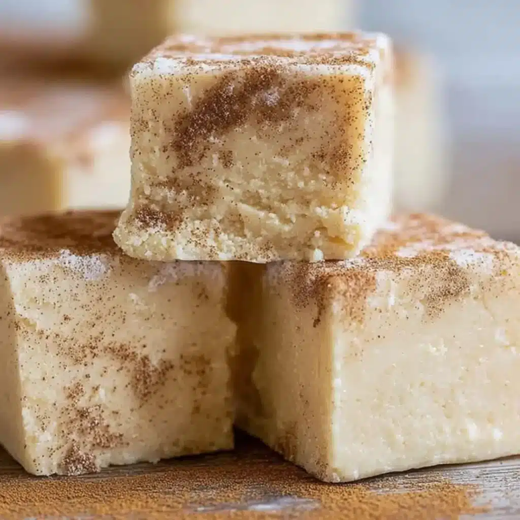 Three square pieces of creamy white snickerdoodle fudge stacked on a wooden surface, dusted with cinnamon sugar.