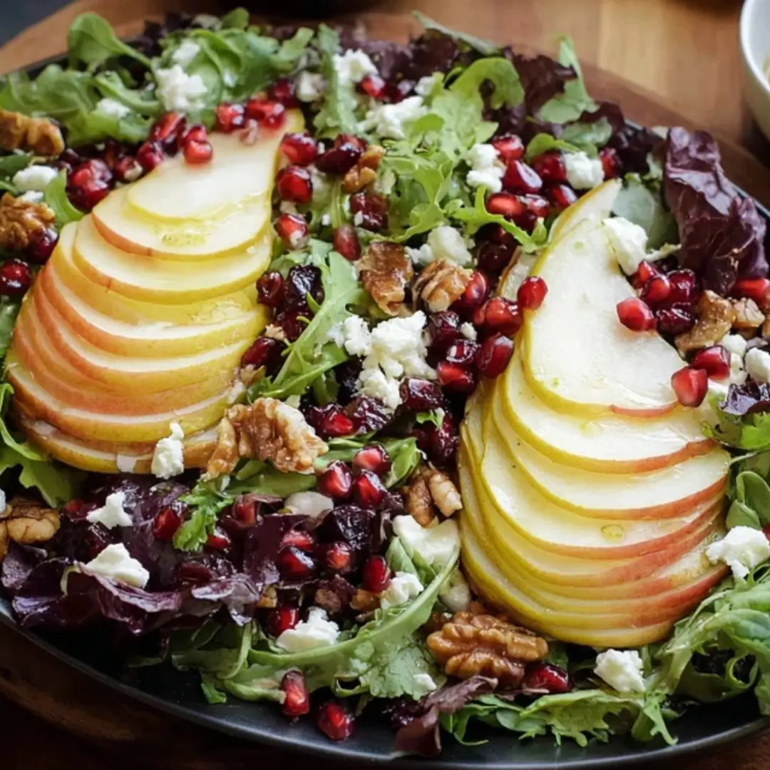 A vibrant Winter Pear Salad featuring thinly sliced ripe pears, crumbled goat cheese, toasted walnuts, and bright red pomegranate seeds over a bed of mixed winter greens.