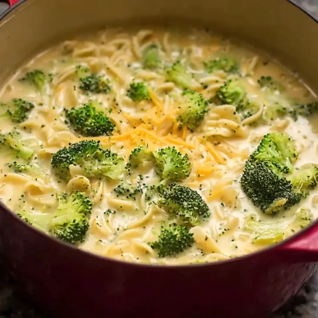 A close-up shot of creamy Broccoli Cheddar Noodle Soup with vibrant green broccoli florets and shredded cheese in a red Dutch oven.