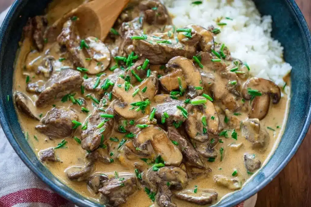A close-up shot of a creamy Beef Stroganoff Recipe in a blue bowl, with tender beef, mushrooms, and chives served with rice.