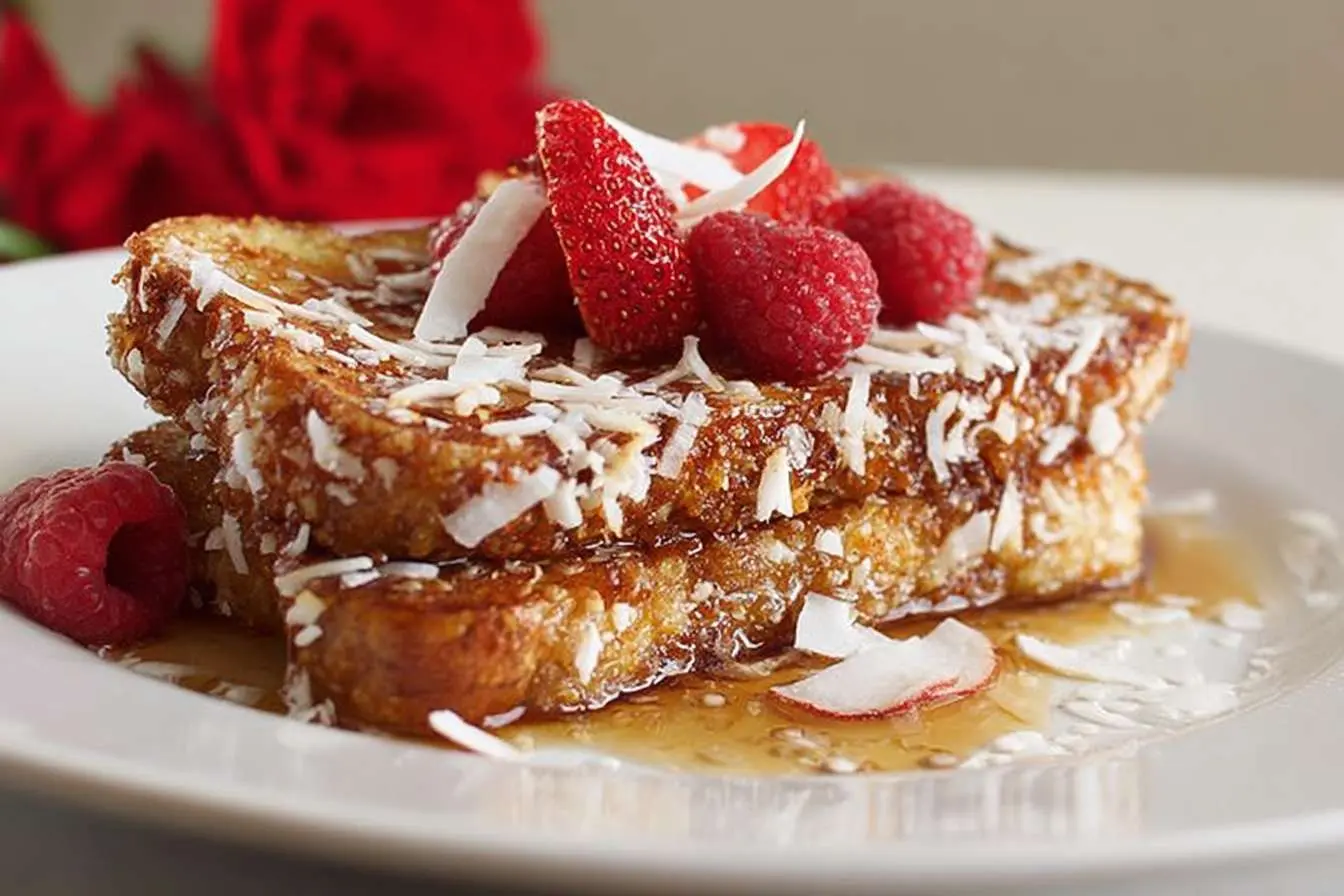 A close-up of stacked Coconut French Toast on a white plate, topped with raspberries, coconut flakes, and drizzled with syrup.