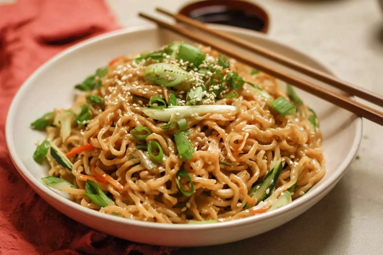 A delicious bowl of creamy peanut noodles topped with scallions and sesame seeds, with chopsticks resting on the side.