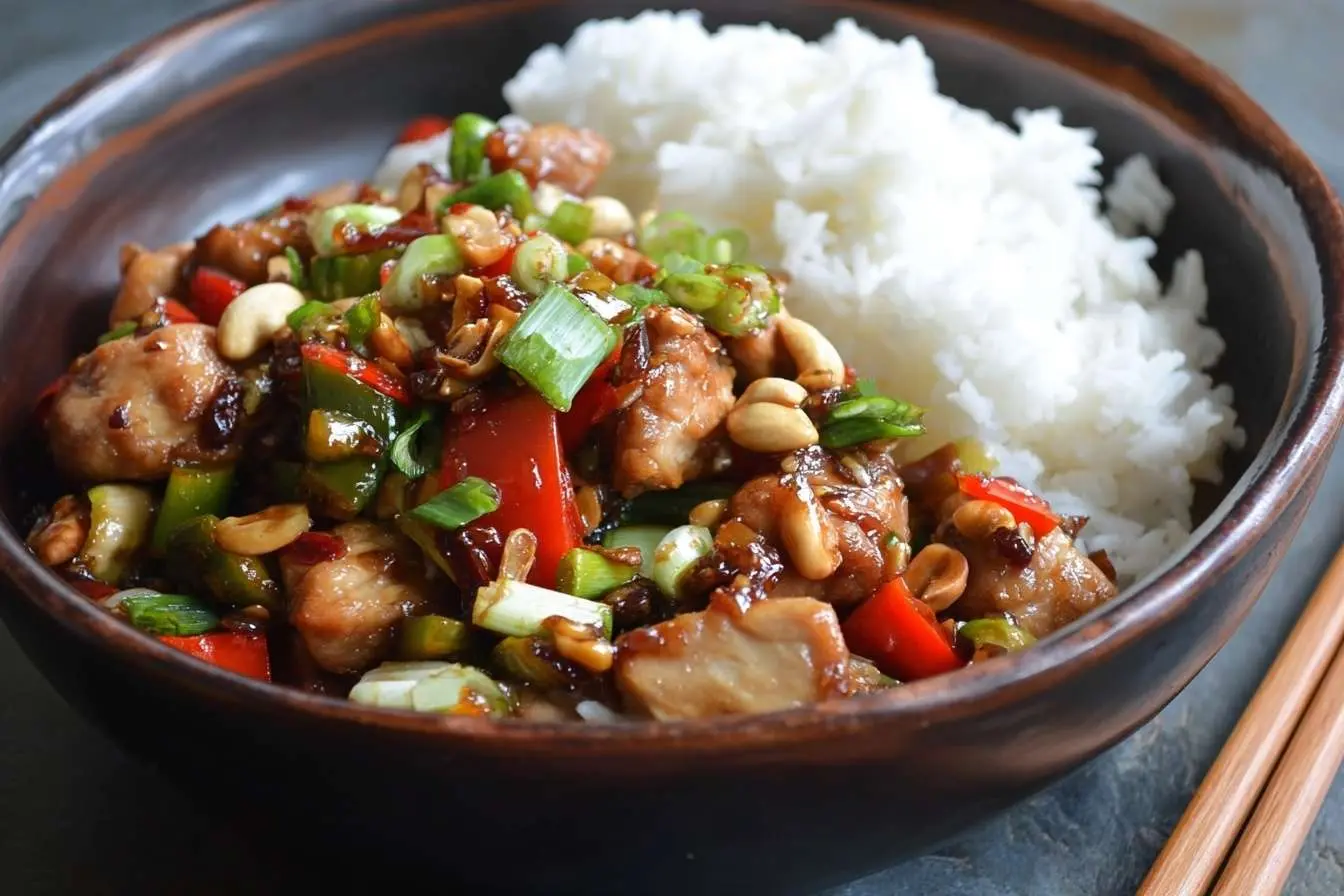 A delicious bowl of Kung Pao Chicken with peanuts, bell peppers, and scallions, served with a side of white steamed rice.