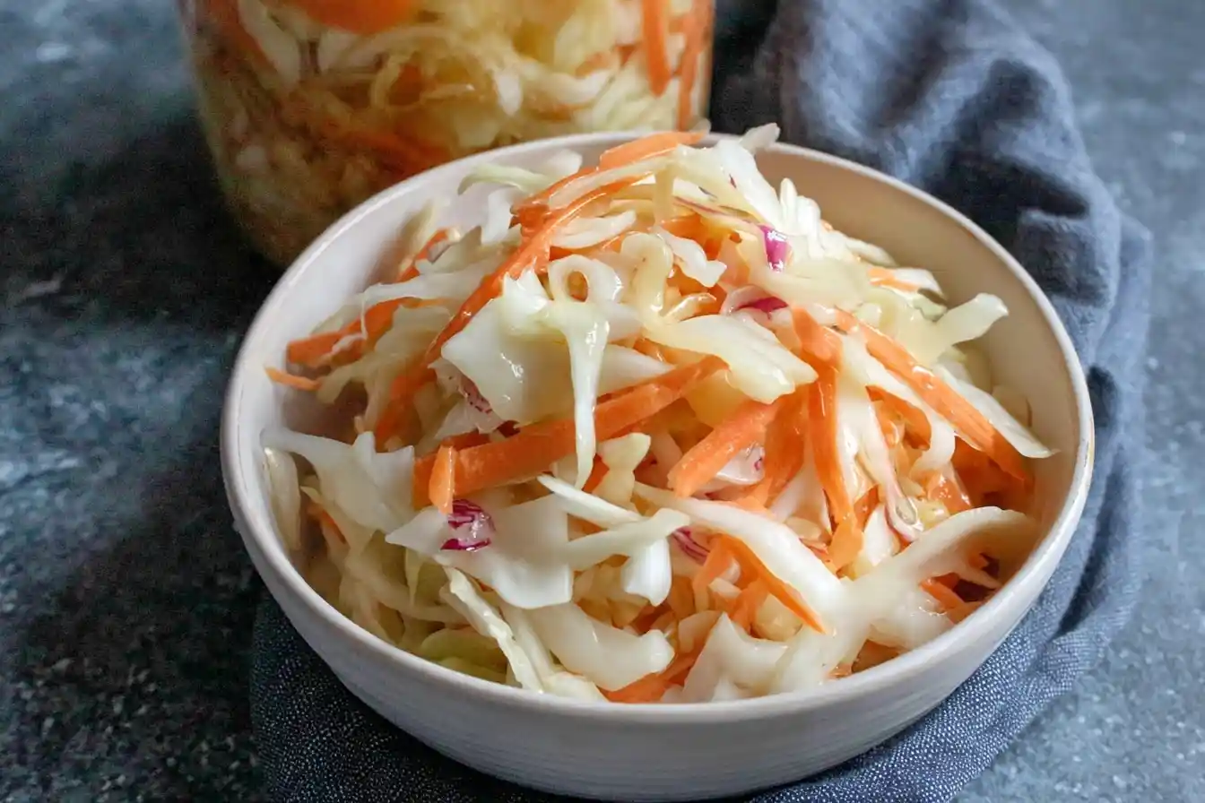 Close-up of a bowl filled with shredded pickled cabbage and carrots next to a glass jar.