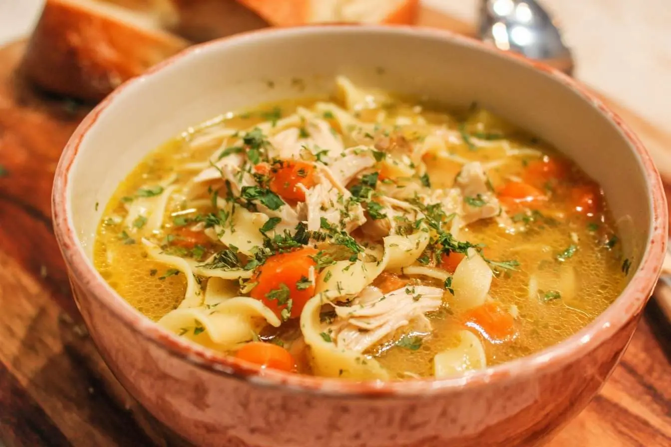 A close-up of a rustic bowl filled with homemade chicken noodle soup, featuring shredded chicken, carrots, and parsley.