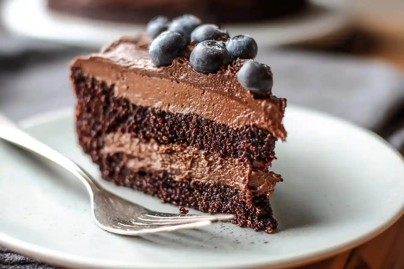 Slice of moist chocolate quinoa cake with thick chocolate frosting and fresh blueberries on a plate with a fork.