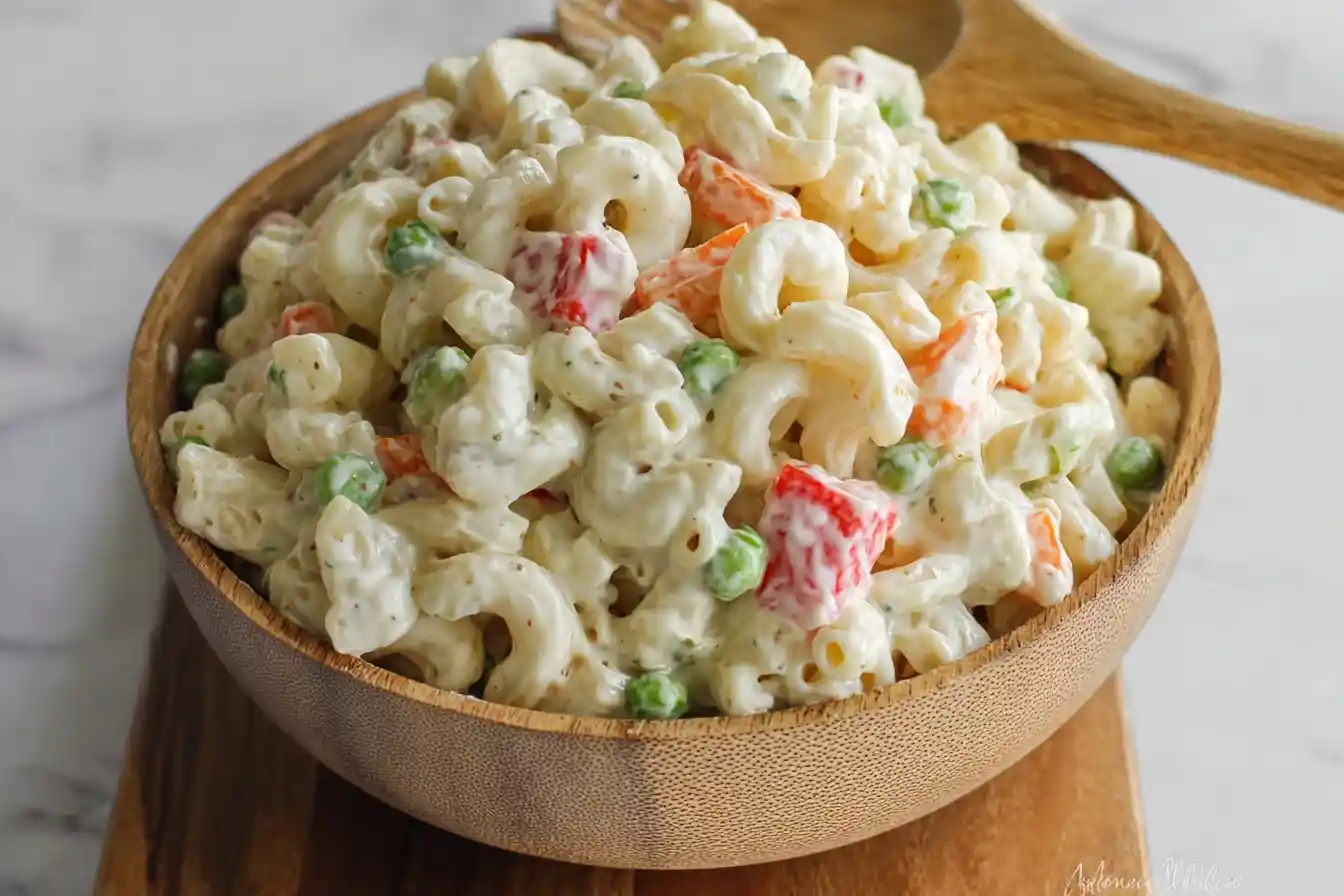 Close-up view of a wooden bowl filled with creamy macaroni salad featuring peas, carrots, and red peppers.