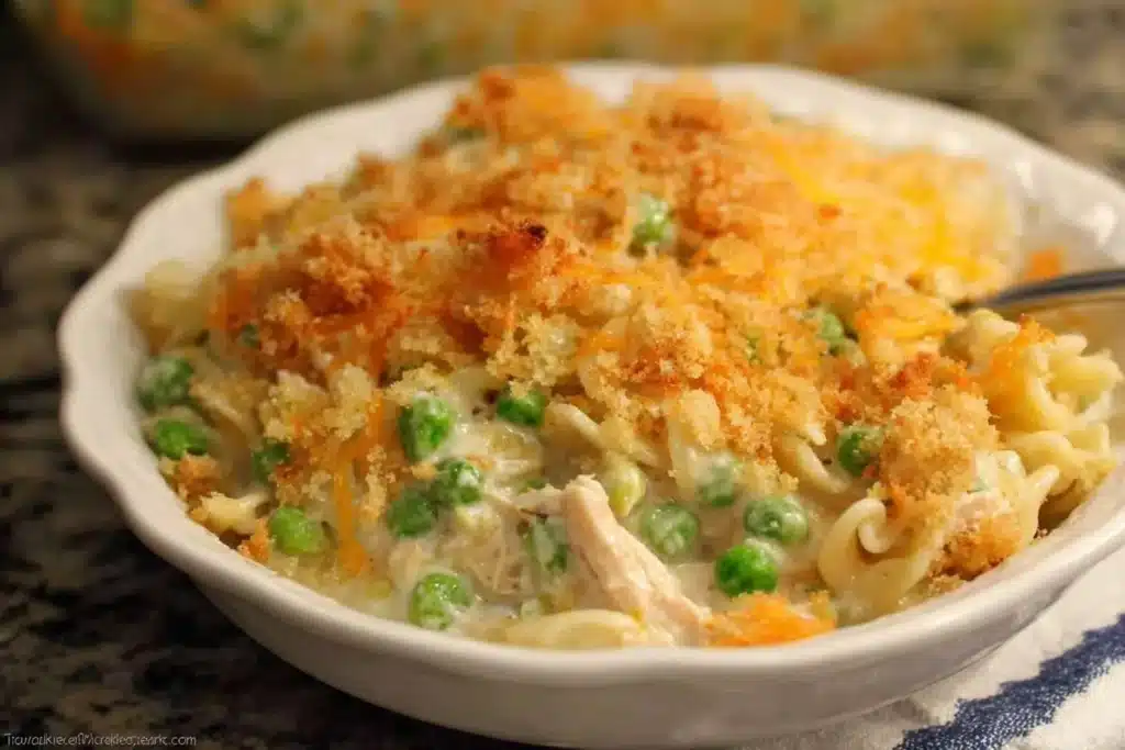 A close-up serving of creamy Tuna Noodle Casserole with peas and a golden breadcrumb topping in a white bowl.