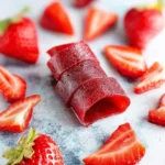 A rolled piece of homemade strawberry fruit leather surrounded by fresh sliced and whole strawberries on a light surface.