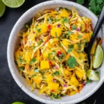 A white bowl filled with vibrant mango slaw, featuring shredded cabbage, fresh mango chunks, cilantro, and lime.