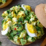 Close-up of a fresh avocado egg salad sandwich on a toasted bun, garnished with fresh dill and red pepper flakes.