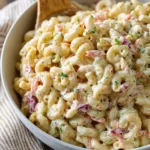 A large white bowl filled with creamy macaroni salad, featuring carrots and red onions, with a wooden spoon.
